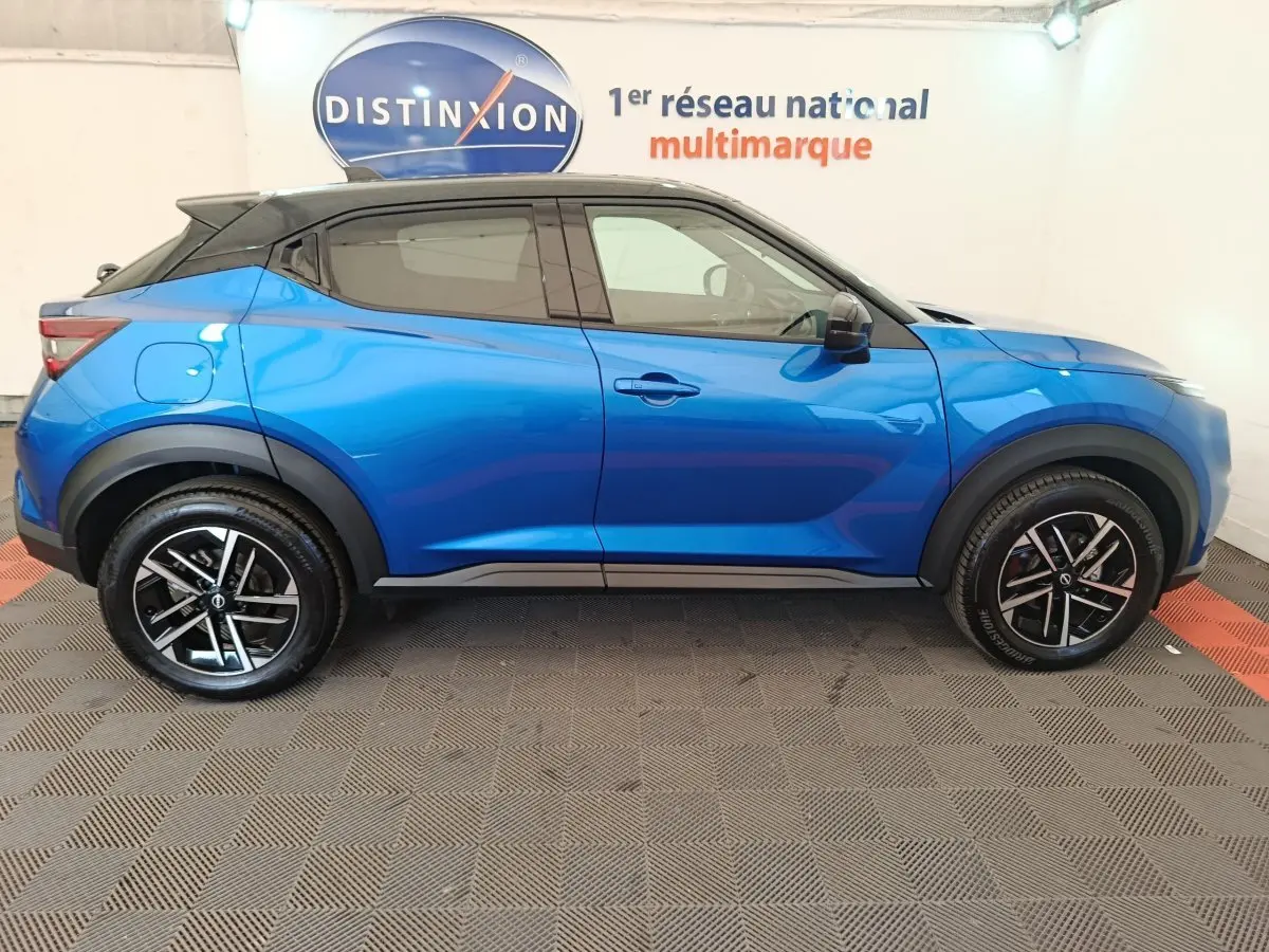 Vue latérale droite d'un Nissan Juke bleu 2025 avec toit noir et jantes alliage noires, en intérieur showroom.