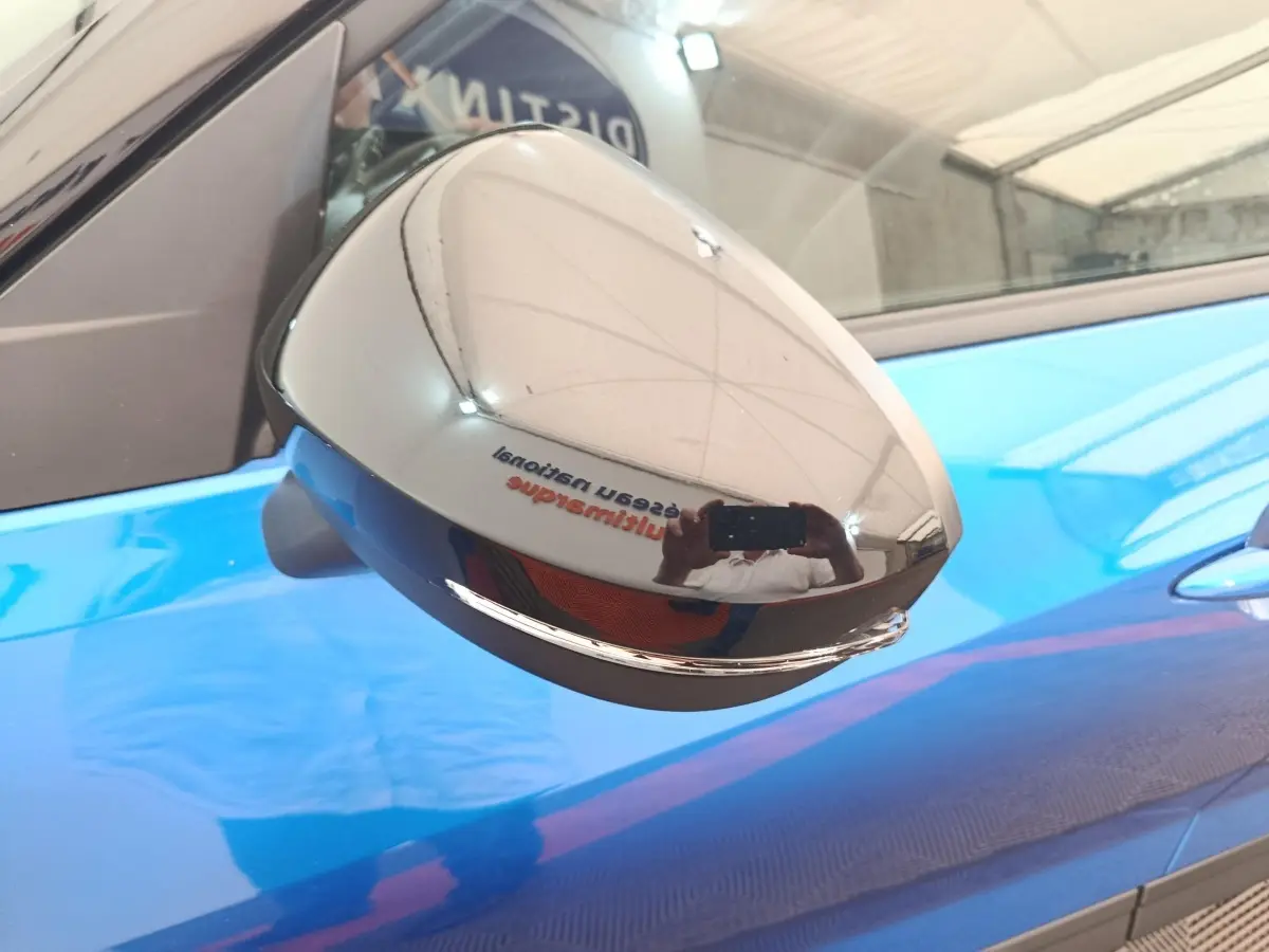 Rétroviseur chromé en gros plan sur le côté gauche d'un Nissan Juke bleu 2025, reflet d'intérieur d'atelier visible.