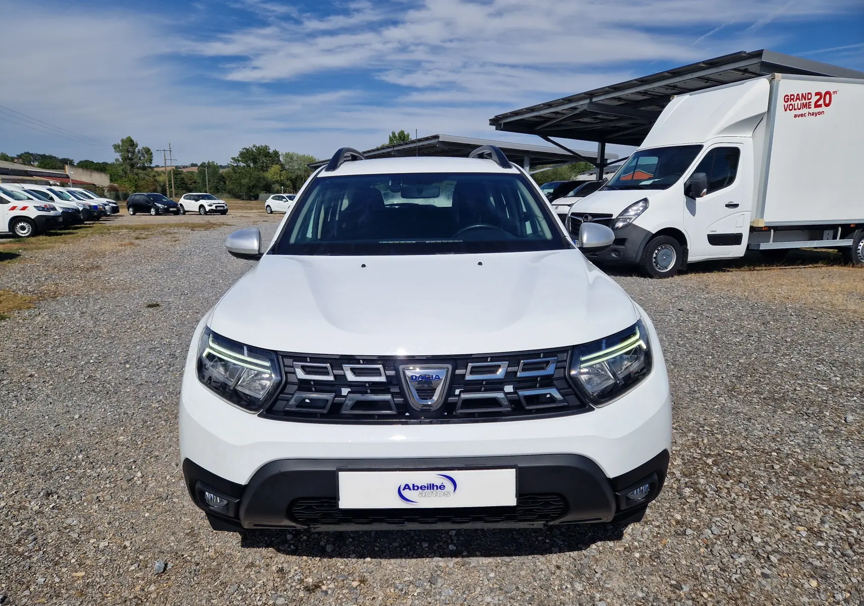 Vue avant d'un Dacia Duster blanc 2023, avec calandre noire et feux à LED allumés, stationné sur un sol gravillonné.