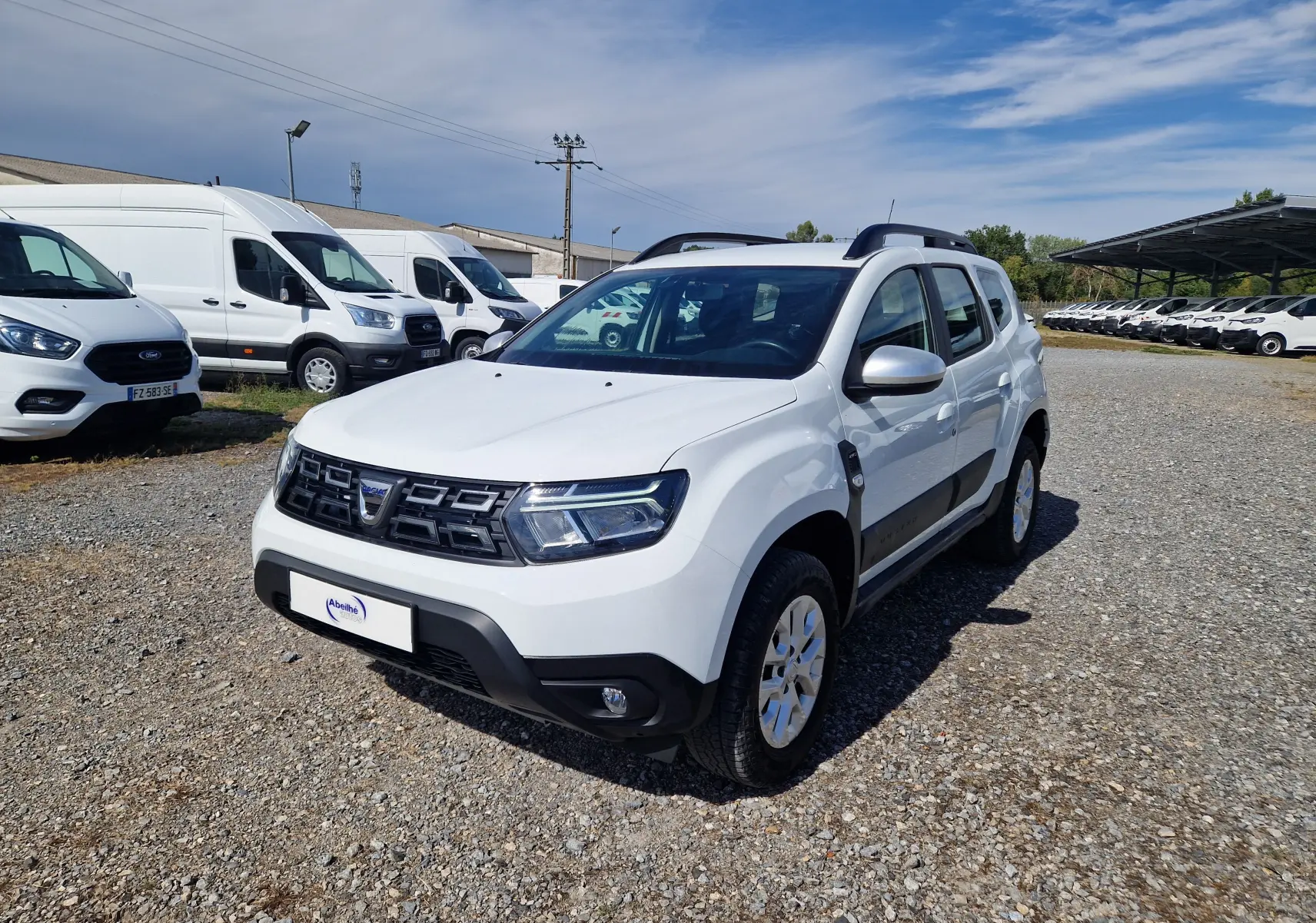 Dacia Duster blanc en 3/4 avant droit, avec calandre noire et barres de toit, sur un sol gravillonné en extérieur.