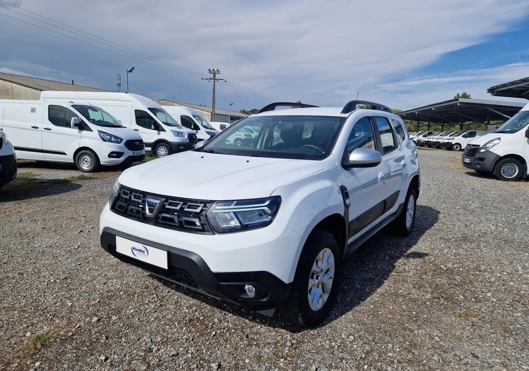 Dacia Duster blanc vue 3/4 avant droit, SUV compact avec protections noires et barres de toit visibles.