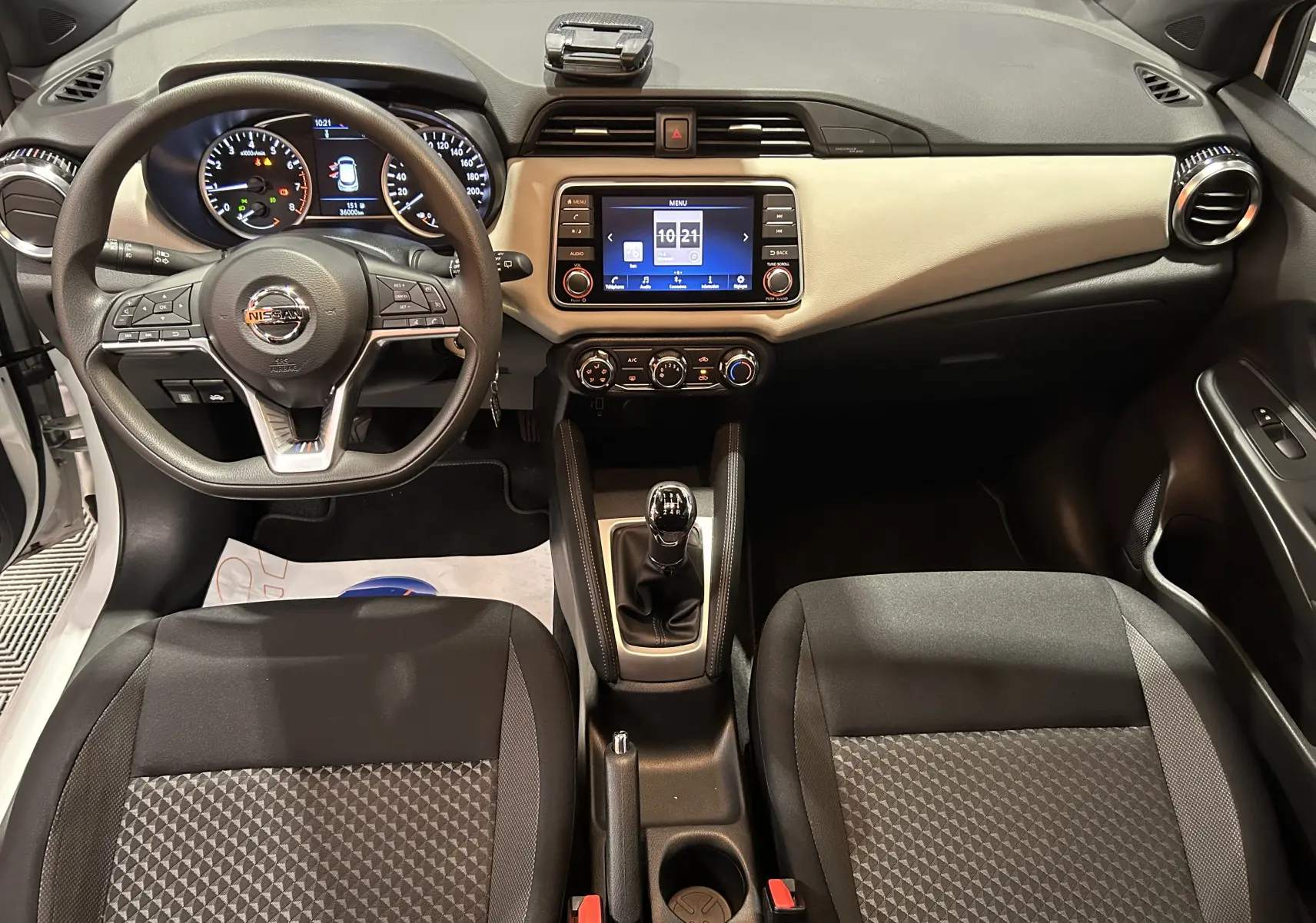 Vue intérieure avant de la Nissan Micra 100ch 2021, tableau de bord beige, volant multifonction et boîte manuelle visible.