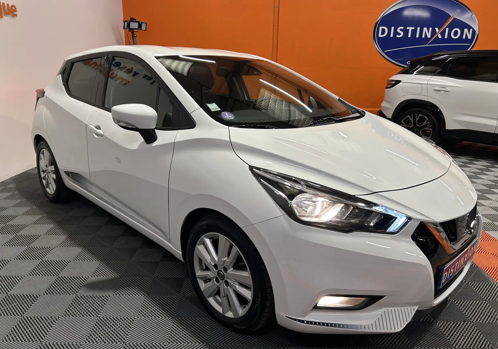 Nissan Micra blanche vue 3/4 avant droit avec phares allumés dans un showroom au sol gris et mur orange.