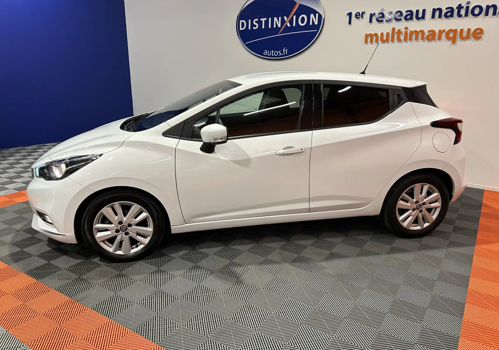 Profil côté gauche d'une Nissan Micra blanche 5 portes de 2021, en intérieur sur sol gris et orange avec logo Distinxion en arrière-plan.