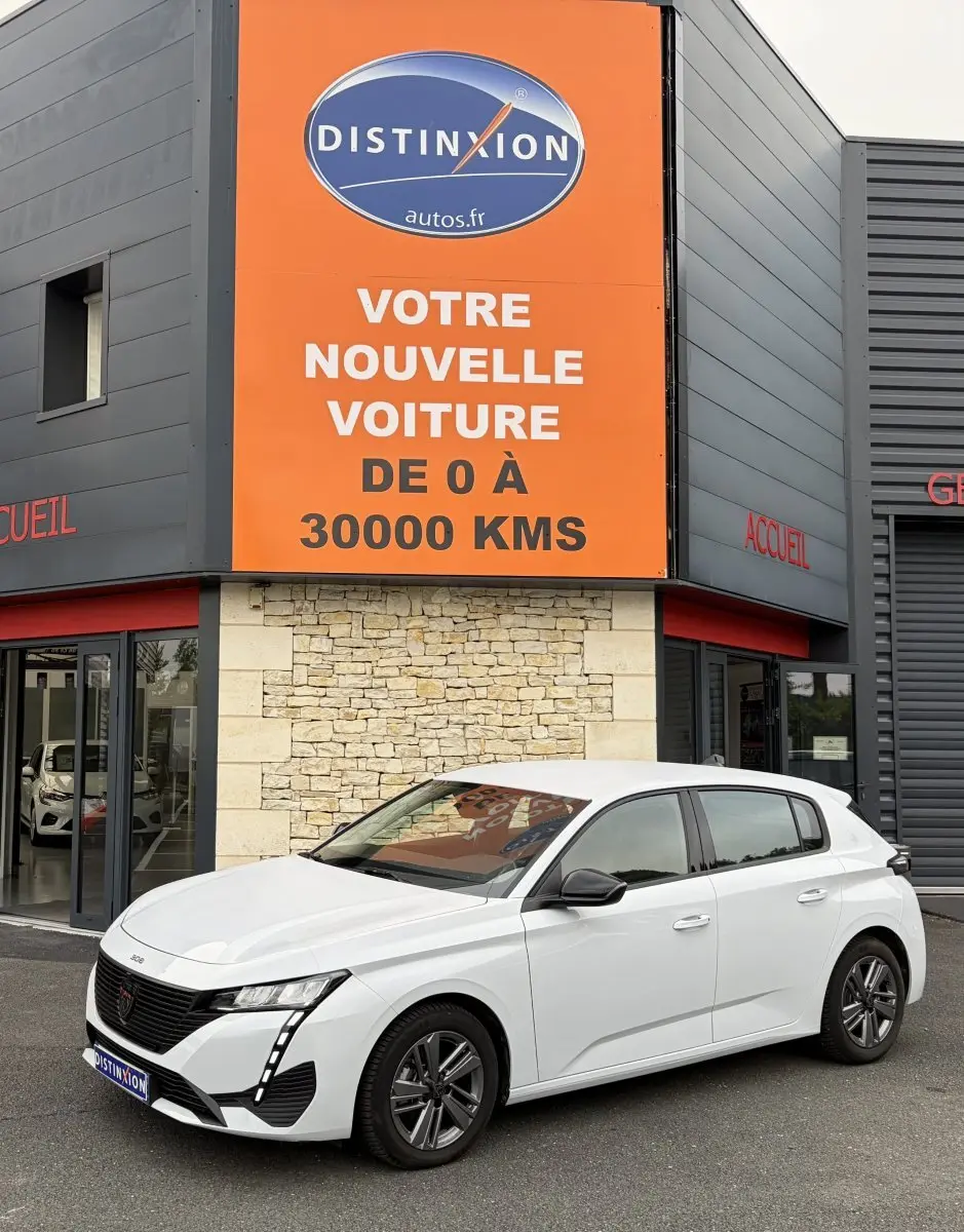 Peugeot 308 blanche vue 3/4 avant droit, avec feux LED verticaux et rétroviseurs noirs devant un bâtiment commercial.