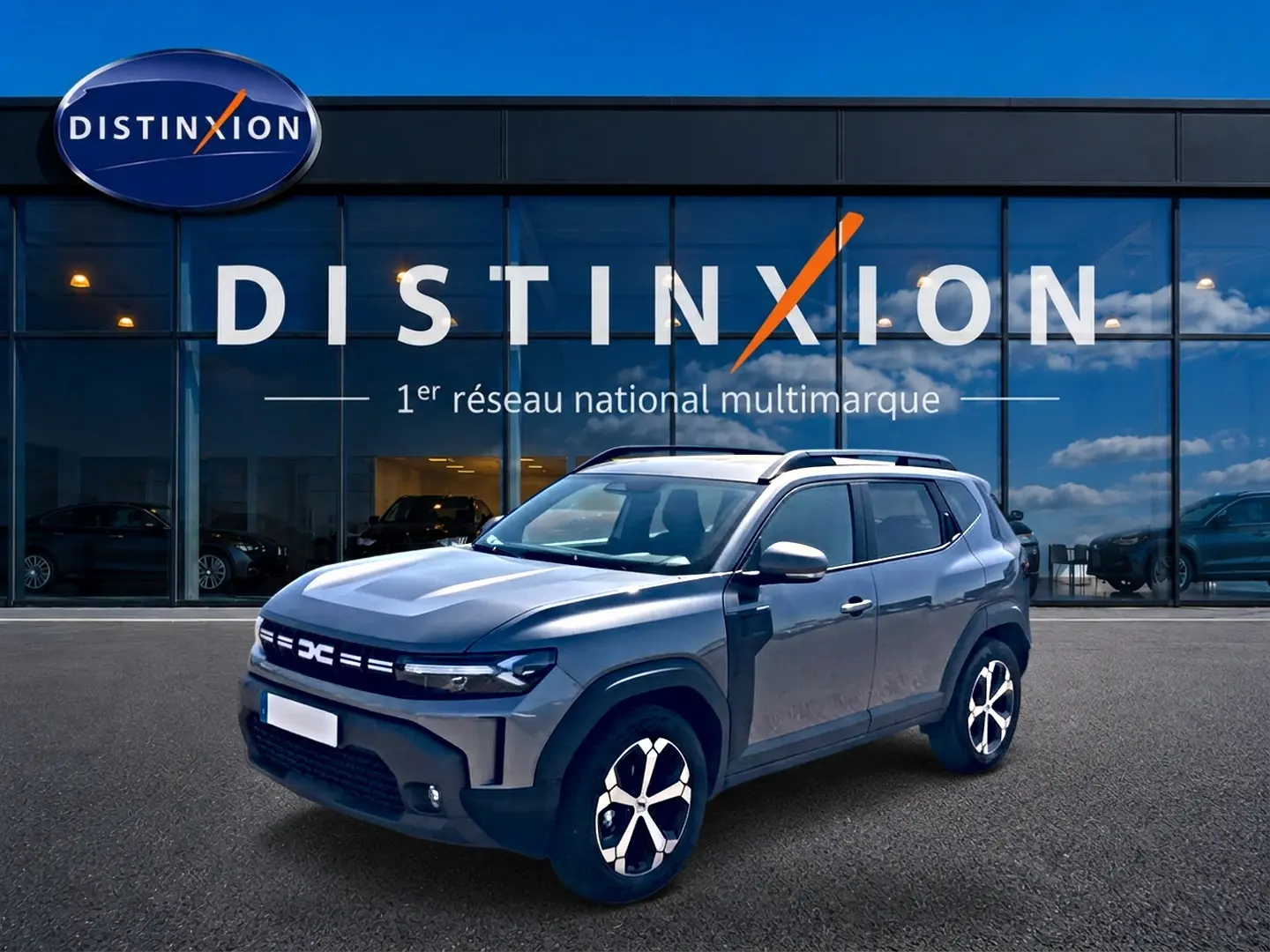 Dacia Duster gris schiste en 3/4 avant droit, avec jantes alu et barres de toit, devant une vitrine Distinxion.