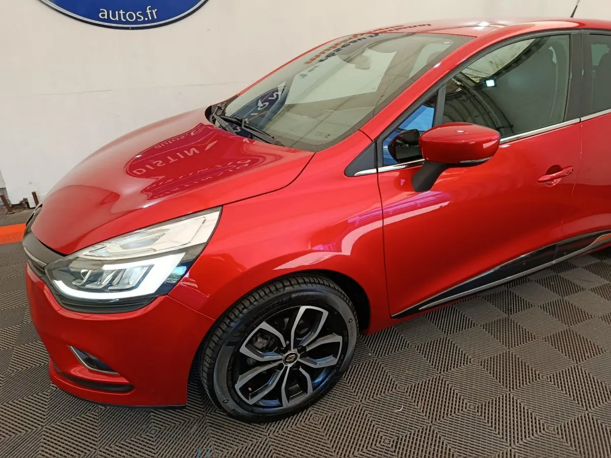 Vue 3/4 avant gauche d'une Renault Clio Intens rouge avec jantes alliage noires et feux LED allumés.