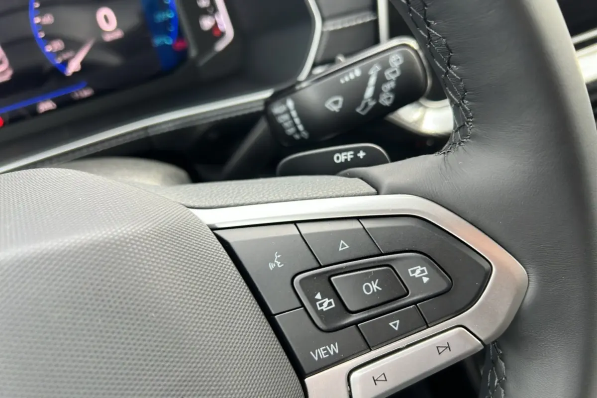 Gros plan sur les commandes multifonctions du volant en cuir du Volkswagen T-Cross gris argent, tableau de bord numérique en arrière-plan.