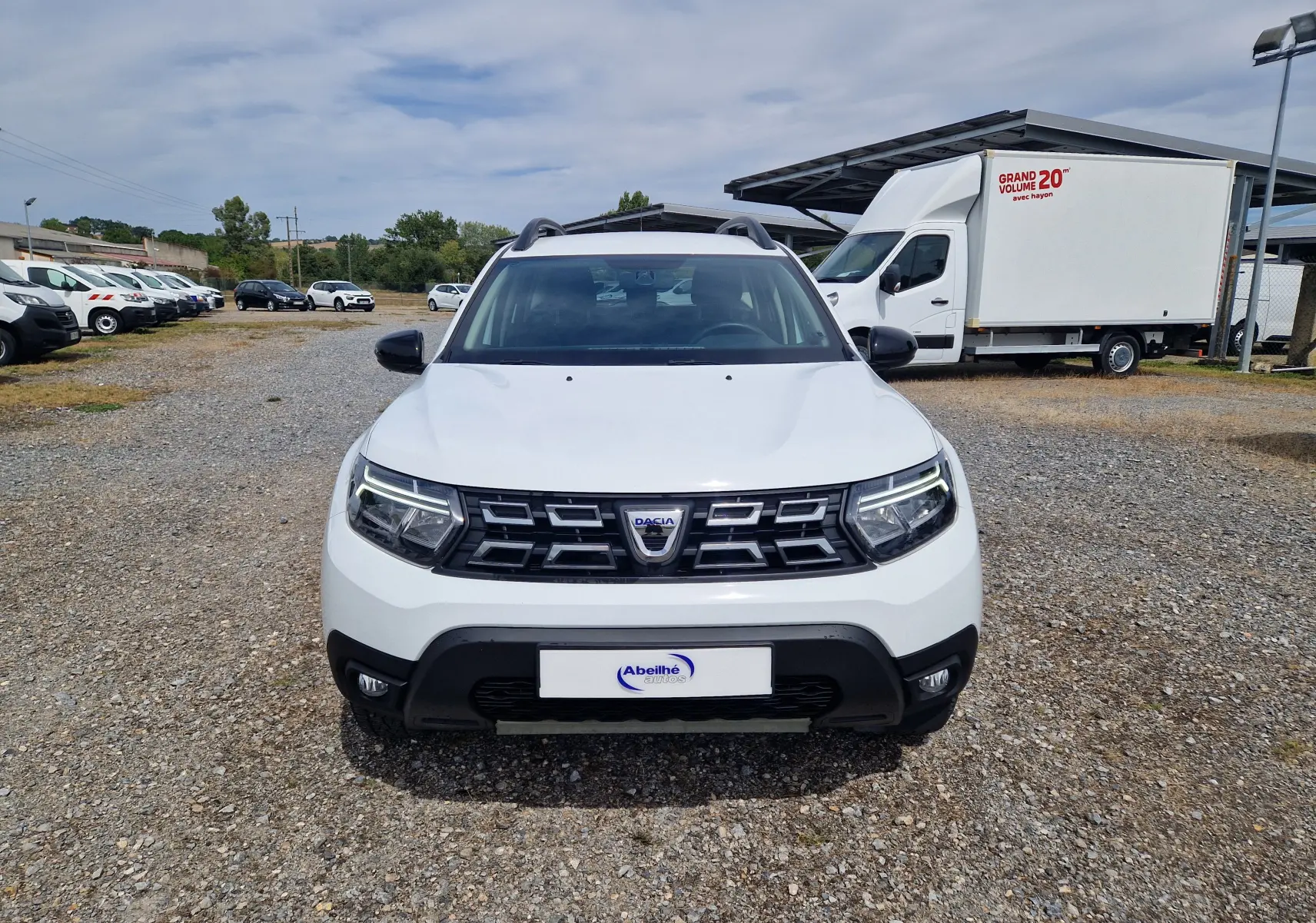 Vue frontale d'un Dacia Duster 4x4 blanc 2022 avec calandre noire et phares LED allumés sur un terrain gravillonné.