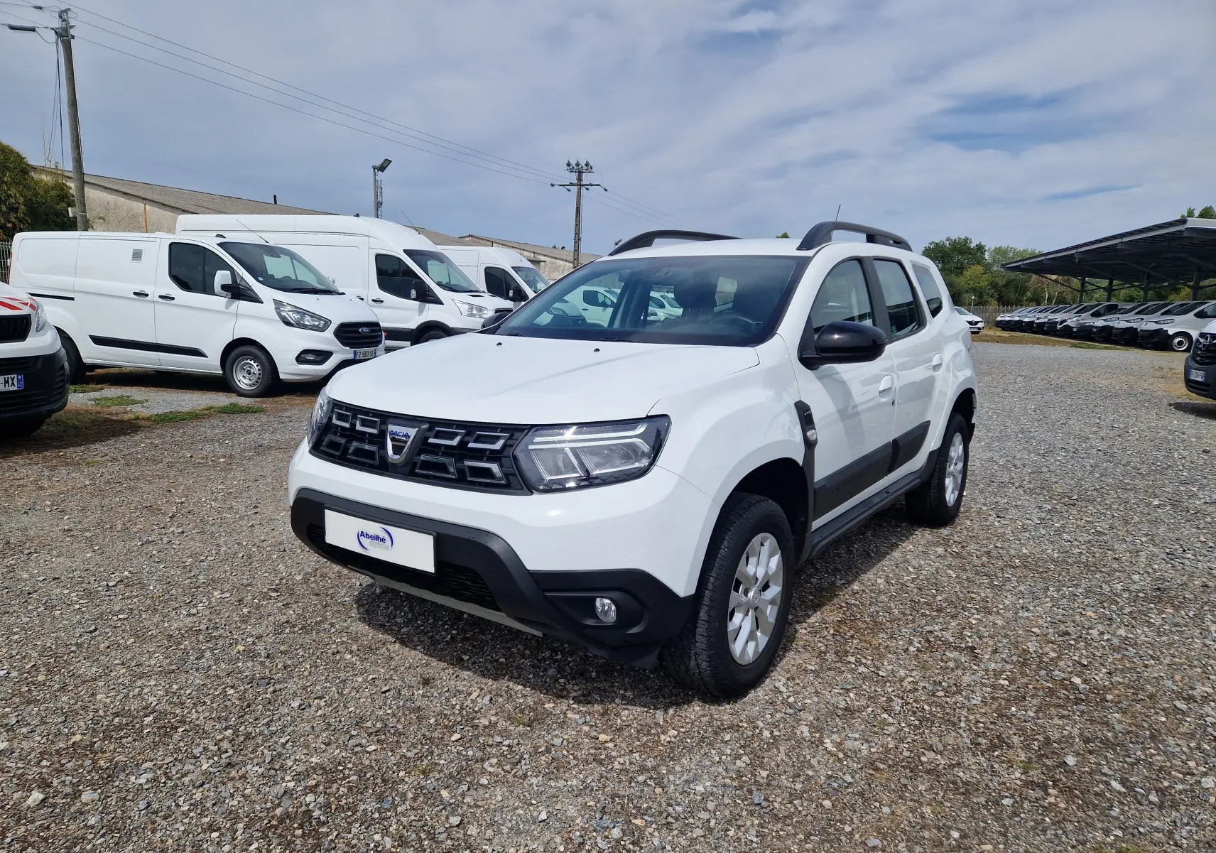 Dacia Duster blanc 4x4 vue 3/4 avant droit, avec protections noires sur les portières et jantes alliage 16 pouces.