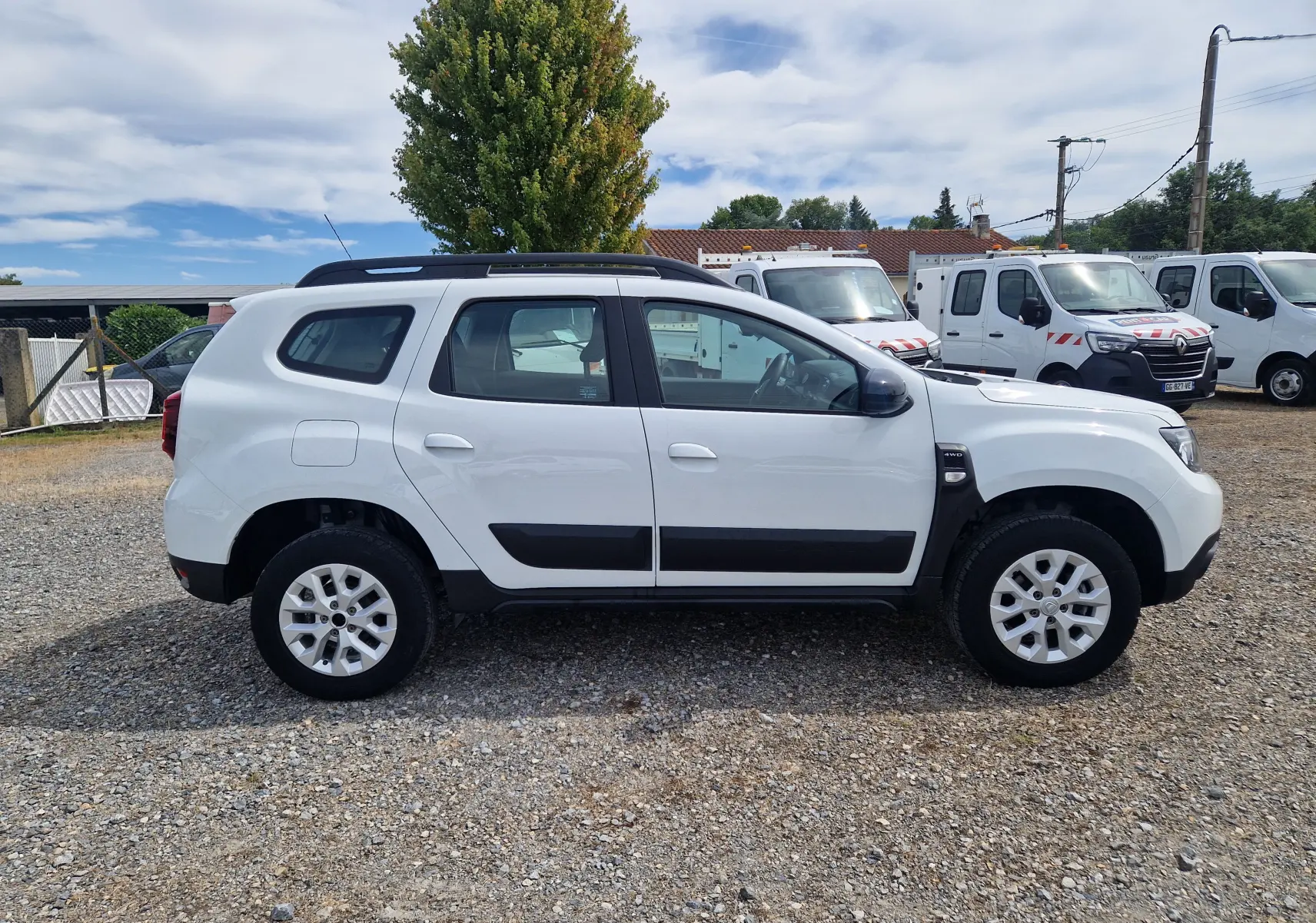 Profil côté gauche d'un Dacia Duster blanc 4x4 avec barres de toit noires, sur un sol gravillonné en extérieur.