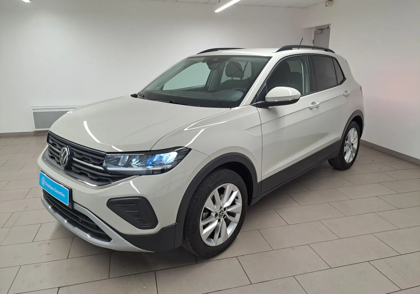 Volkswagen T-Cross Gris Ascot en vue 3/4 avant droit, avec phares allumés et jantes alliage visibles.