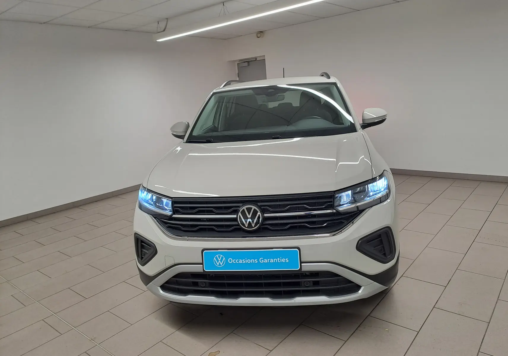 Vue avant d'un Volkswagen T-Cross 2024 gris Ascot avec phares allumés dans un showroom blanc.
