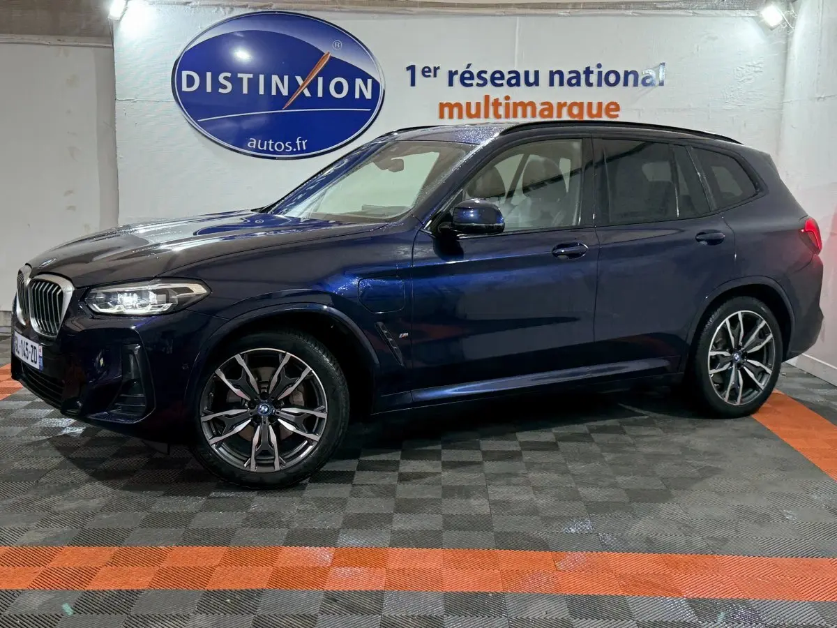 BMW X3 xDrive30e bleu en vue de profil côté gauche dans un showroom avec éclairage intérieur et logo Distinxion en arrière-plan