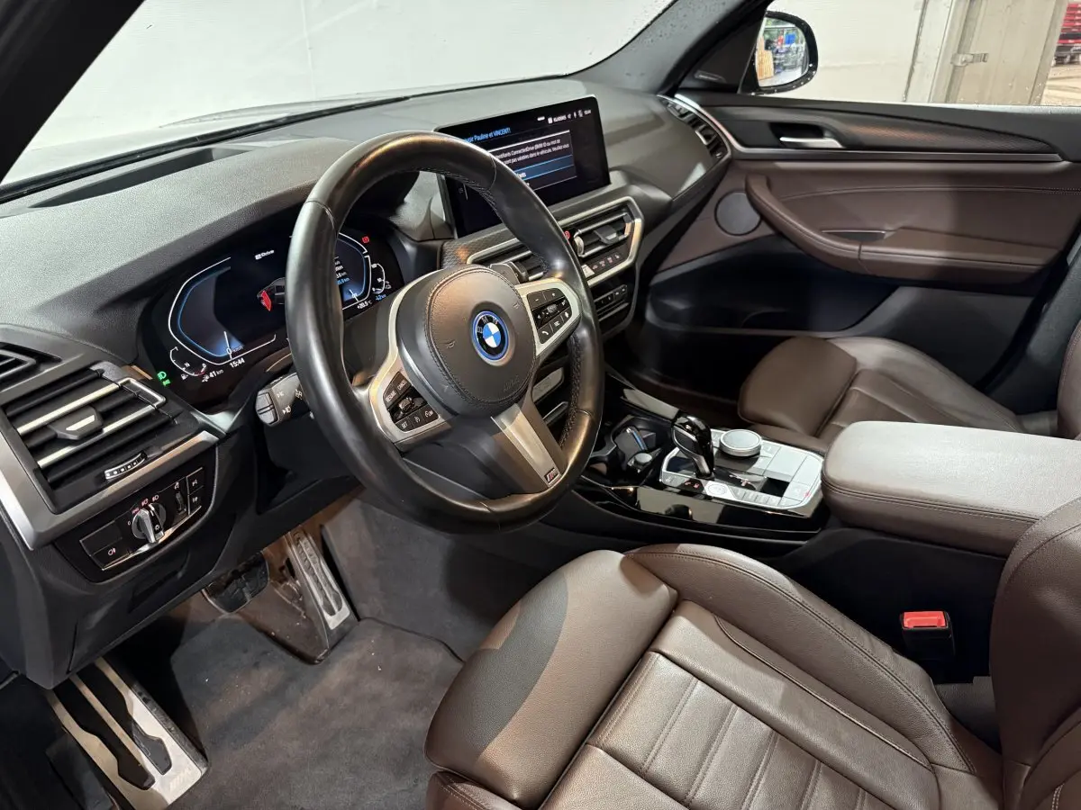 Intérieur BMW X3 xDrive30e 2023 vu côté conducteur, volant cuir, écran tactile et sièges marron cuir.