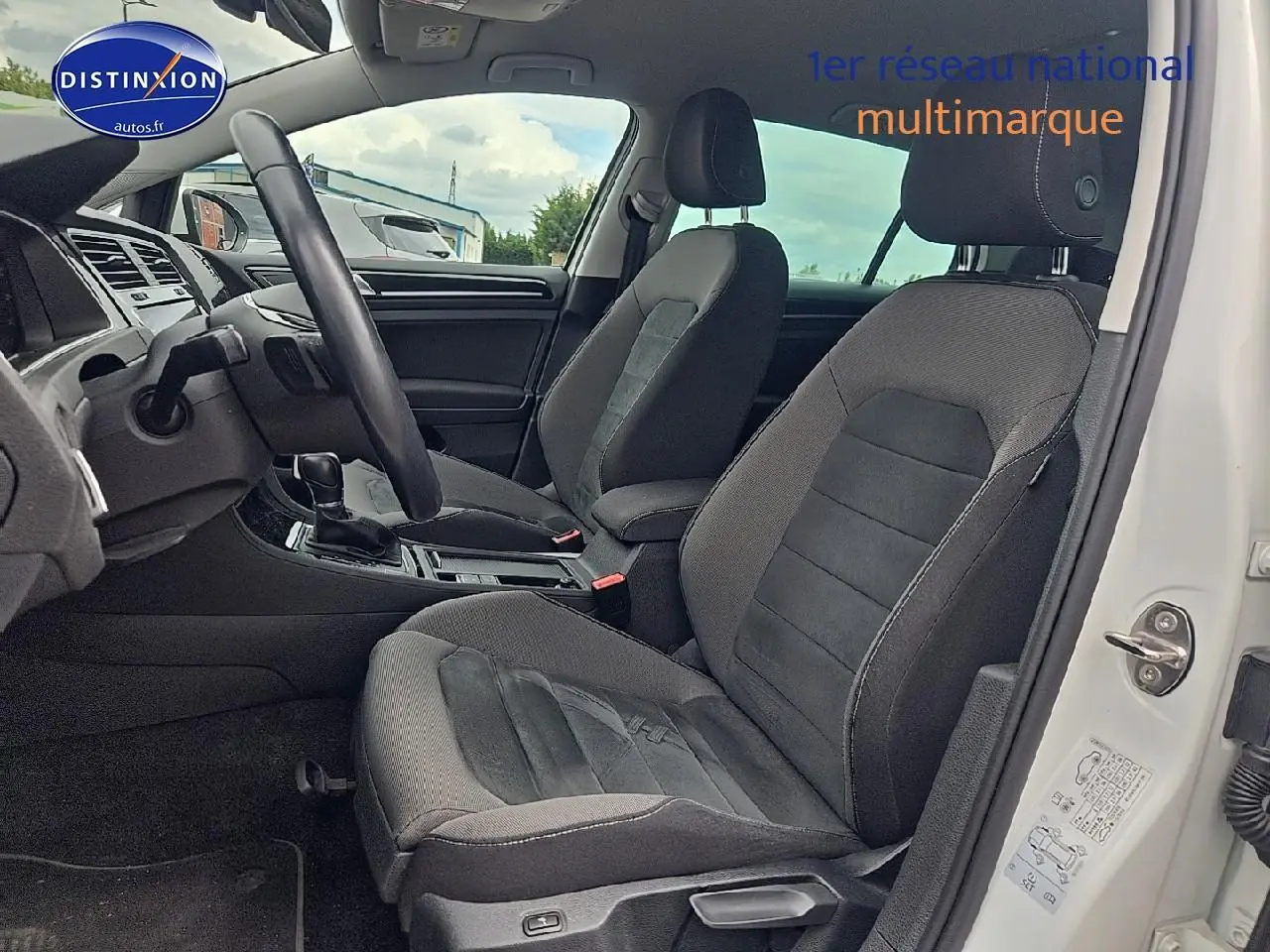 Vue intérieure côté conducteur de la Volkswagen Golf blanche 2019, montrant les sièges avant en tissu gris et la console centrale.