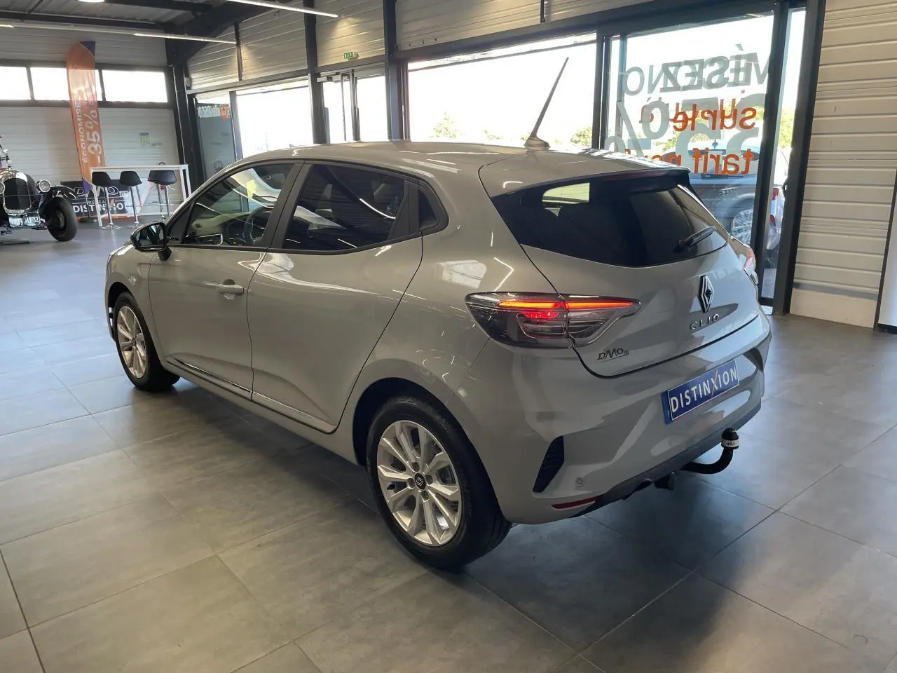 Renault Clio 1.5 Blue dCi 2025 gris Rafale vue 3/4 arrière droit en intérieur avec feux arrière allumés.