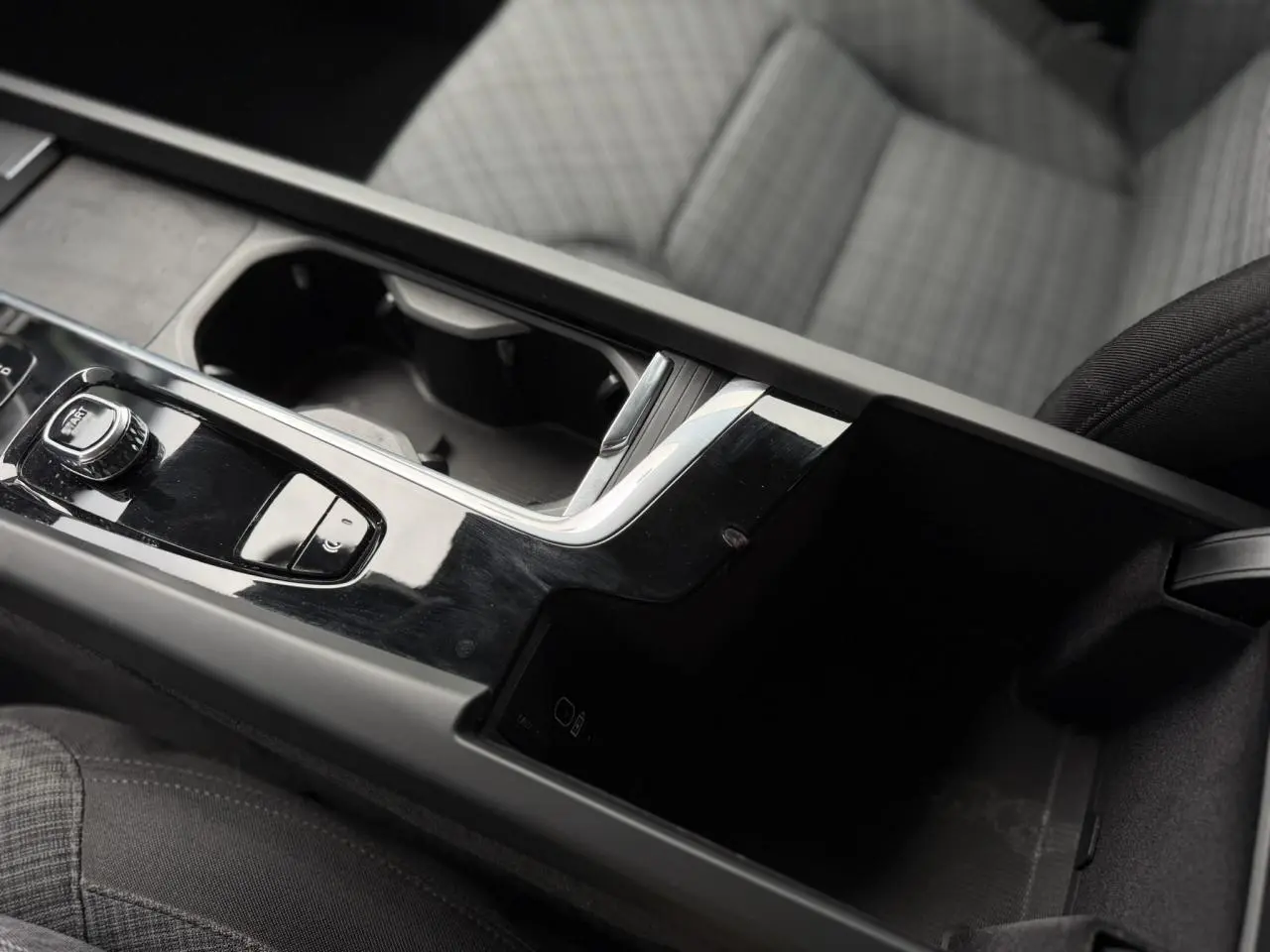 Gros plan sur la console centrale noire brillante de la Volvo S60 2024 avec accoudoir et sièges gris à motifs.