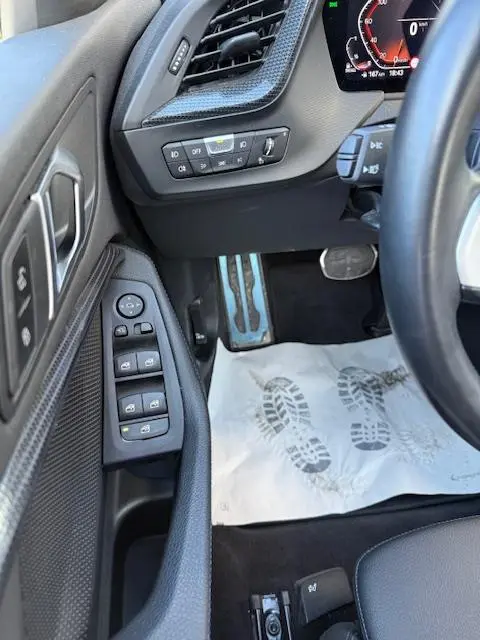 Vue intérieure côté conducteur de la BMW Série 1 118d Edition Sport 2024, montrant les commandes de porte et pédales en aluminium.