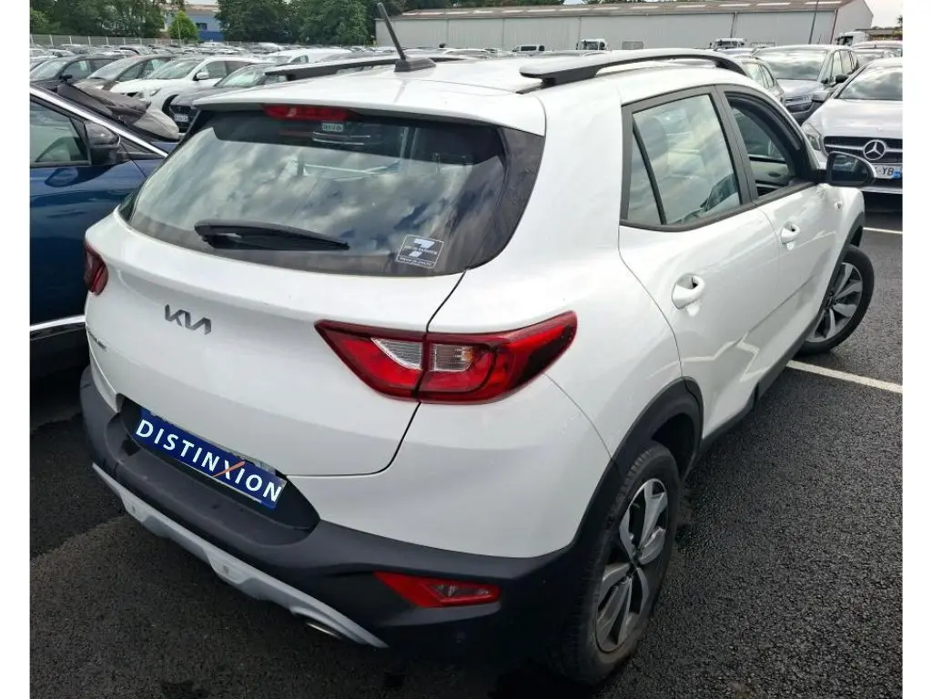 Vue 3/4 arrière droite d’un KIA STONIC blanc 2023 avec feux arrière rouges et toit avec barres de toit noires.