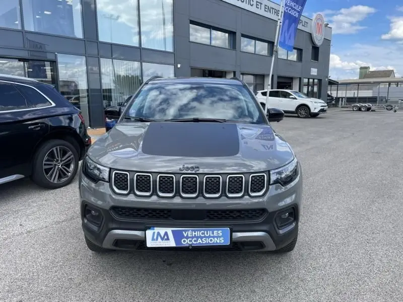 Vue de face d'un Jeep Compass gris 2023 avec capot noir mat et calandre distinctive en sept fentes, stationné devant un concessionnaire.