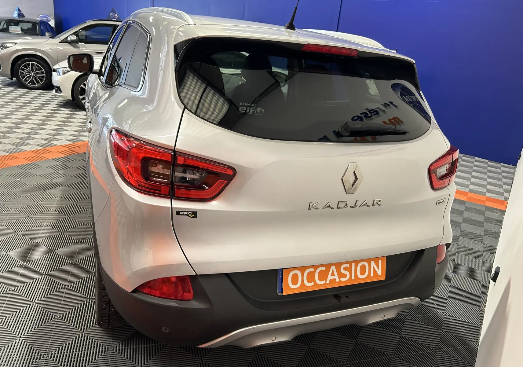 Vue 3/4 arrière droite d'un Renault Kadjar gris avec feux arrière LED et plaque orange "OCCASION" en intérieur showroom.