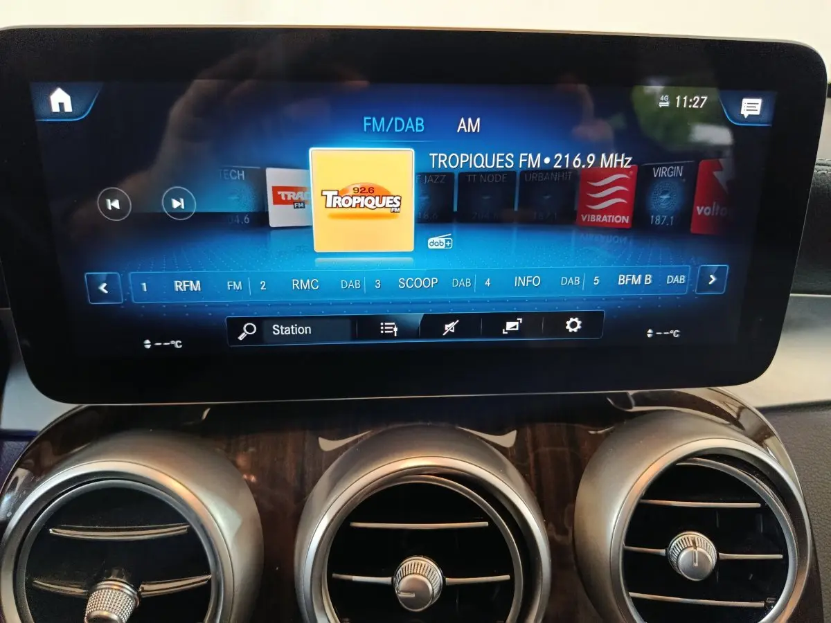Écran tactile central allumé affichant une radio FM, avec aérateurs ronds et tableau de bord bois dans un Mercedes GLC noir.