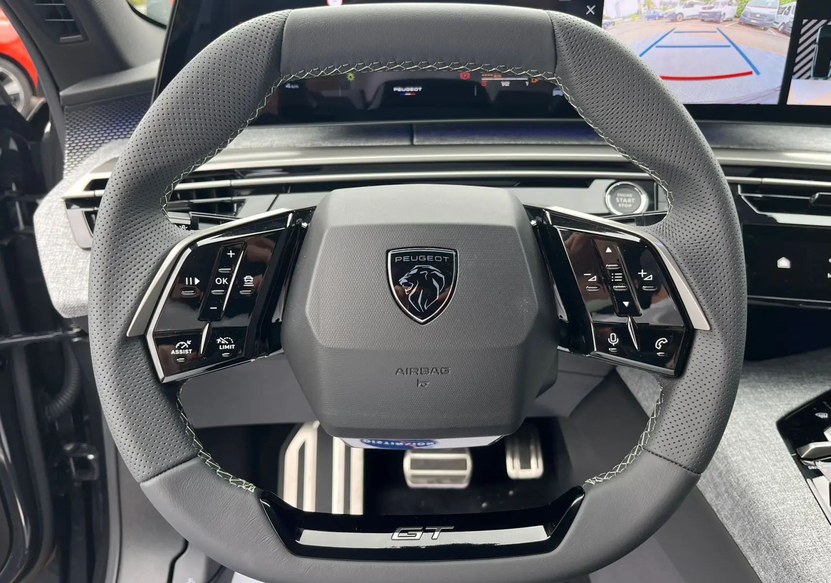 Gros plan sur le volant cuir perforé gris avec logo Peugeot et inscription GT, tableau de bord numérique en arrière-plan.