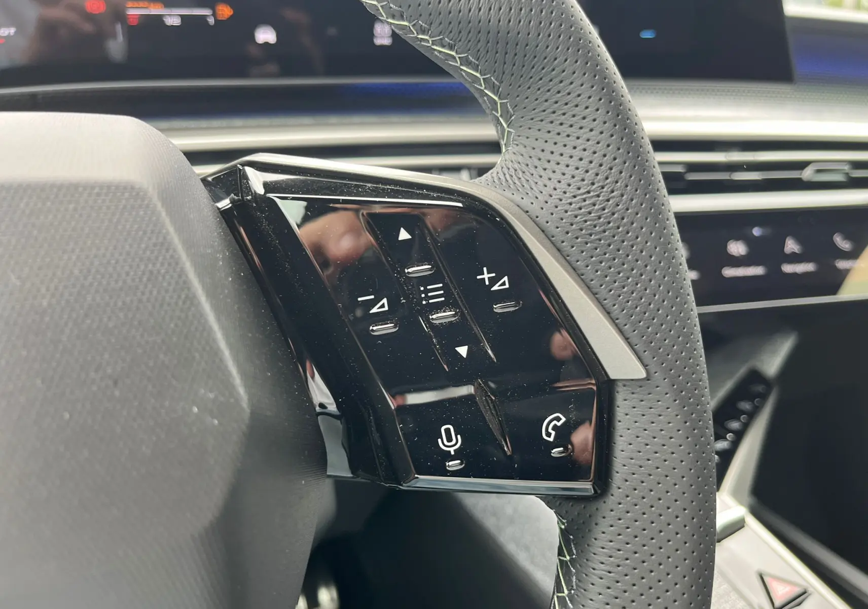 Gros plan sur les commandes gauche du volant en cuir perforé du Peugeot 3008 gris titane, avec tableau de bord numérique en arrière-plan.