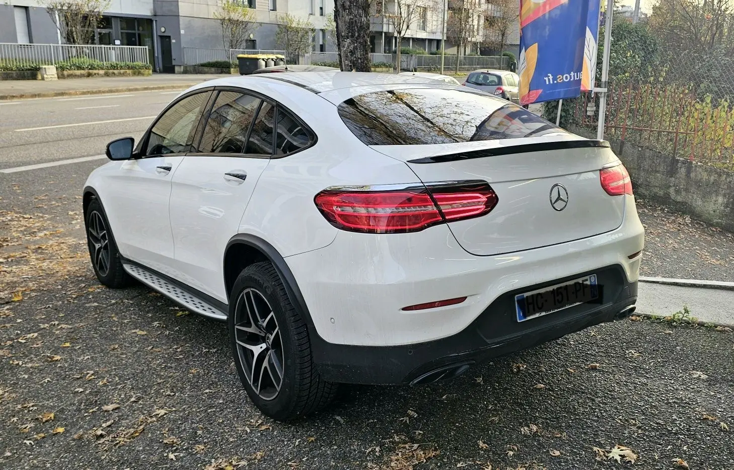 Vue 3/4 arrière droite d'un Mercedes GLC Coupé blanc avec jantes noires et vitres teintées sur parking urbain.