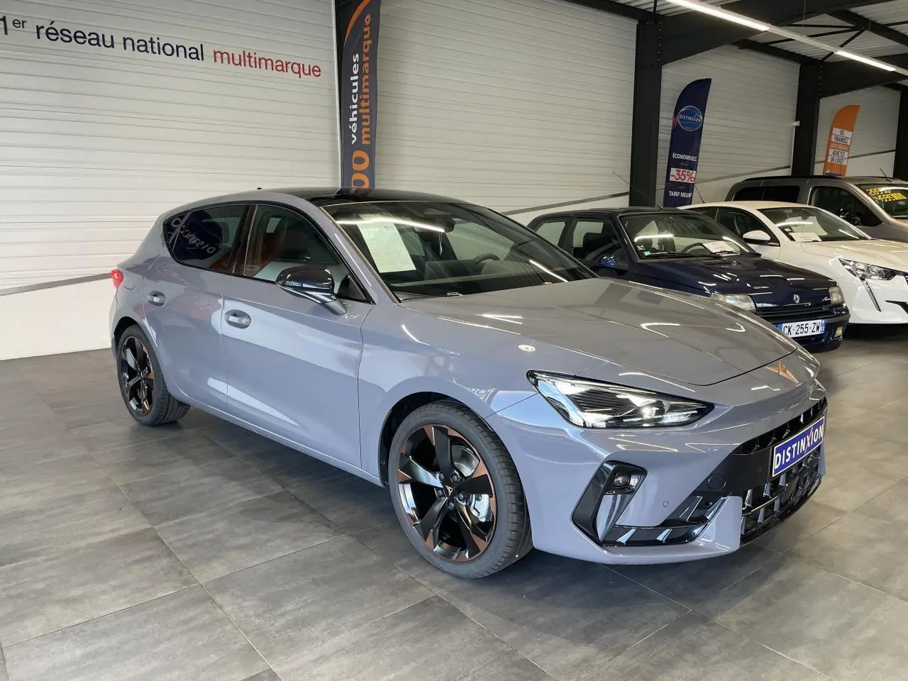 Vue 3/4 avant de la CUPRA Leon gris avec jantes cuivrées et toit ouvrant panoramique en intérieur showroom.