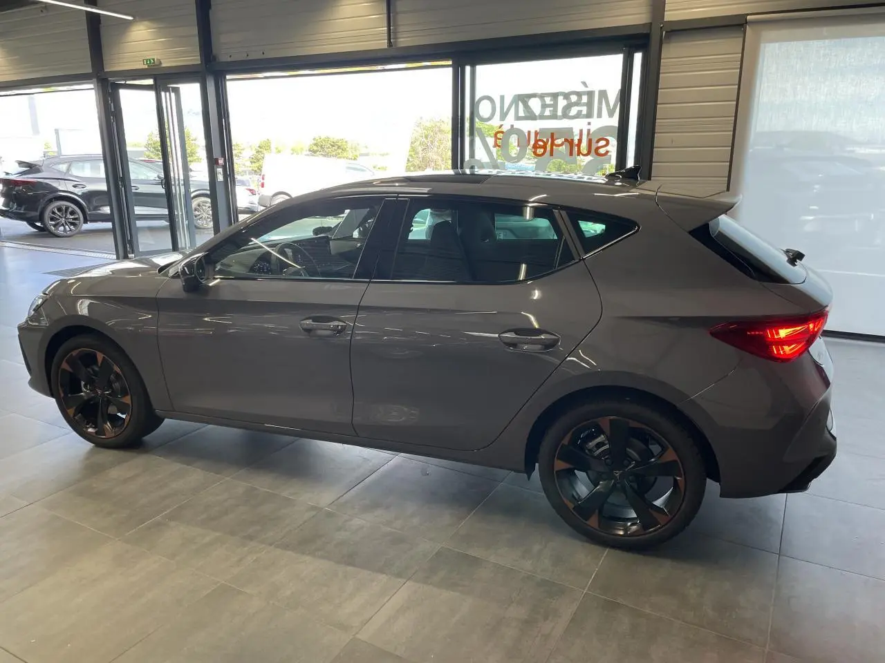 Vue de profil côté gauche d'une CUPRA Leon gris magnétique avec jantes alliage 18'' cuivrées en showroom.