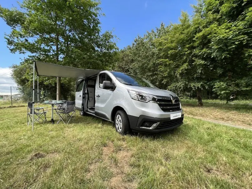 Renault Trafic Fourgon gris en 3/4 avant droit avec porte latérale ouverte et auvent déployé sur herbe.