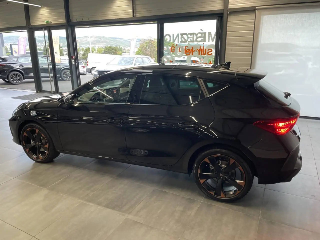 Vue de profil côté gauche d'une CUPRA Leon noire avec jantes alliage 18'' cuivrées en intérieur showroom.
