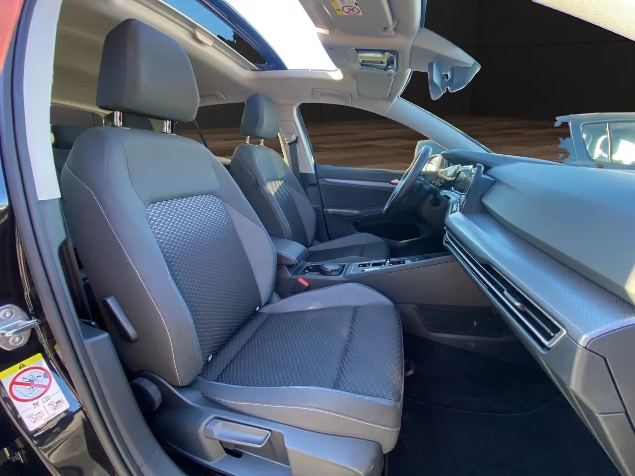 Vue intérieure côté gauche de la Volkswagen Golf VIII noire, mettant en valeur les sièges tissu noirs et le toit panoramique.