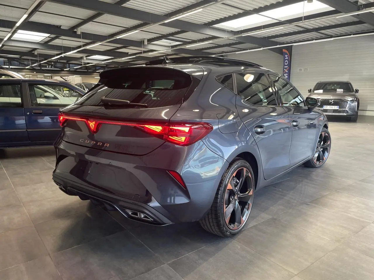 Vue 3/4 arrière droite d'une CUPRA Leon gris magnétique avec jantes alliage 18'' cuivrées et feux arrière Tri-LED allumés.