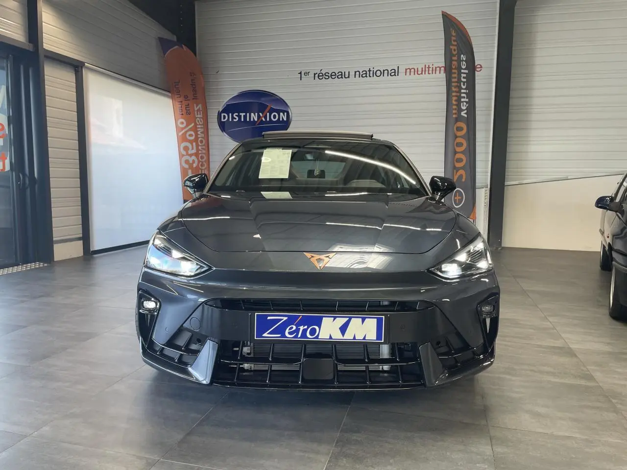 Vue frontale d'une CUPRA Leon gris magnétique avec logo cuivré et phares LED allumés en showroom.