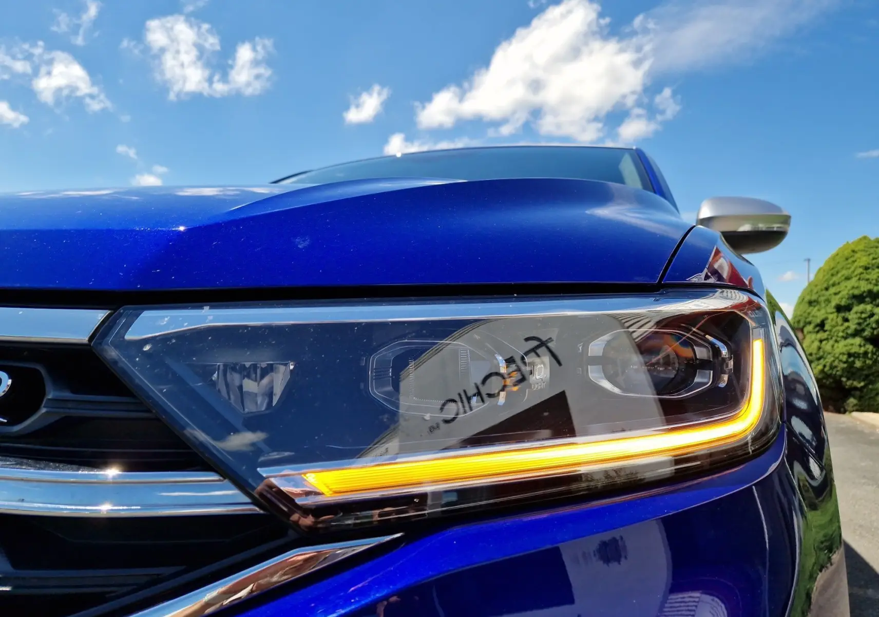 Gros plan sur le phare avant droit du Volkswagen T-Roc bleu Lapiz avec éclairage LED allumé sous un ciel bleu.
