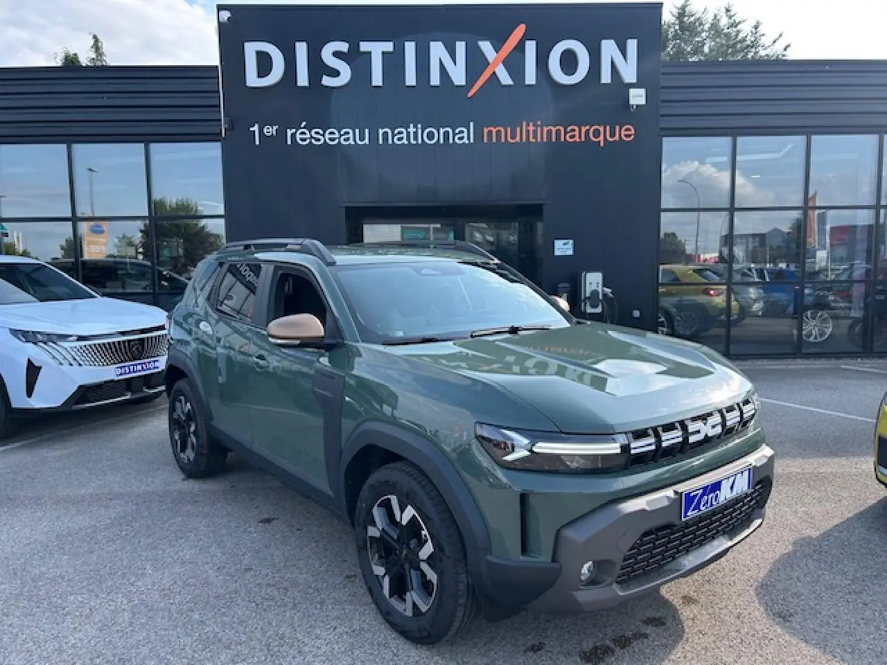 Vue 3/4 avant droit d'un Dacia Duster 2025 kaki dusty avec jantes alliage et barres de toit noires devant un showroom Distinxion.