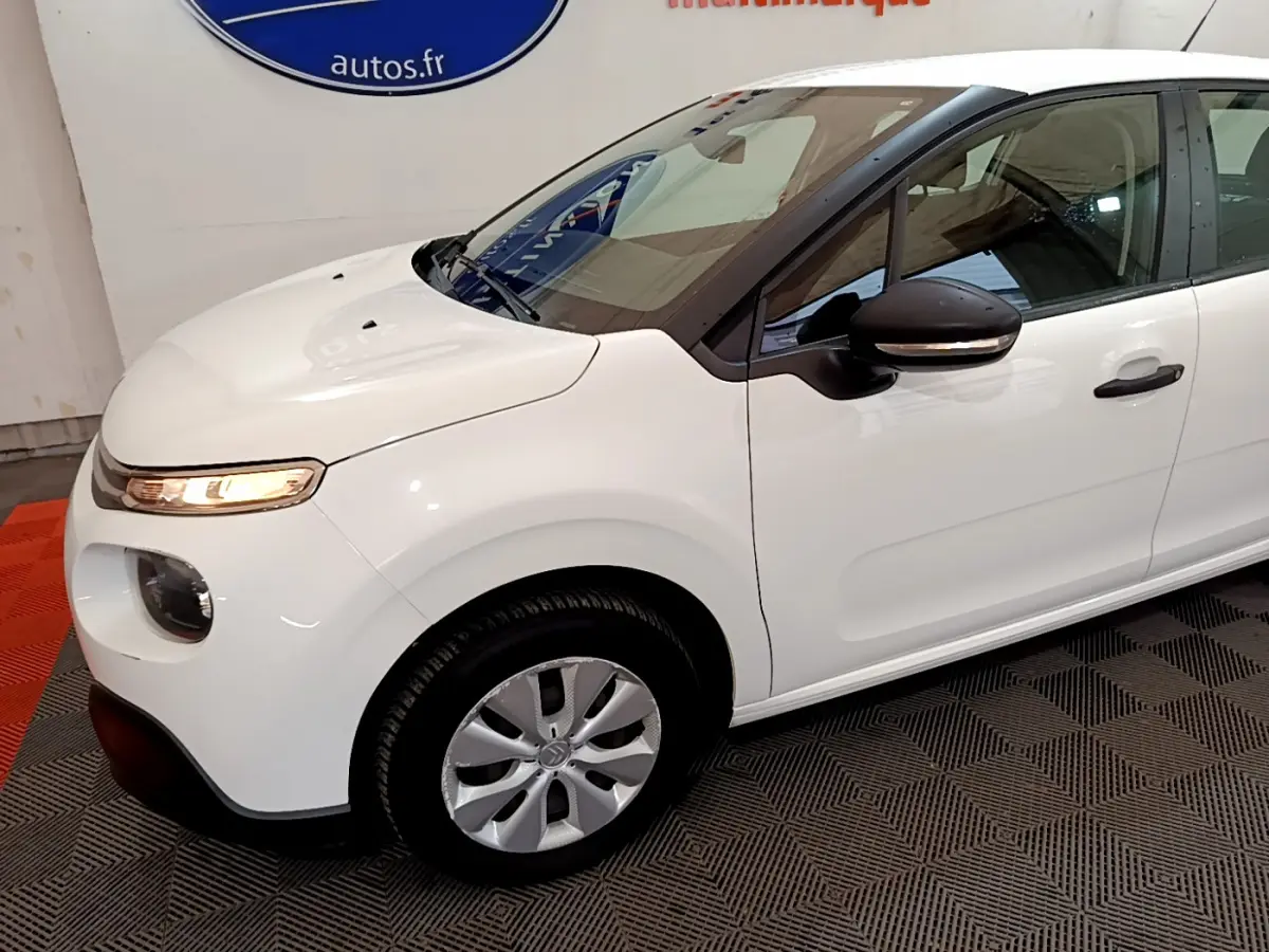 Vue 3/4 avant gauche d'une Citroën C3 blanche avec rétroviseur noir et feux avant allumés en intérieur.