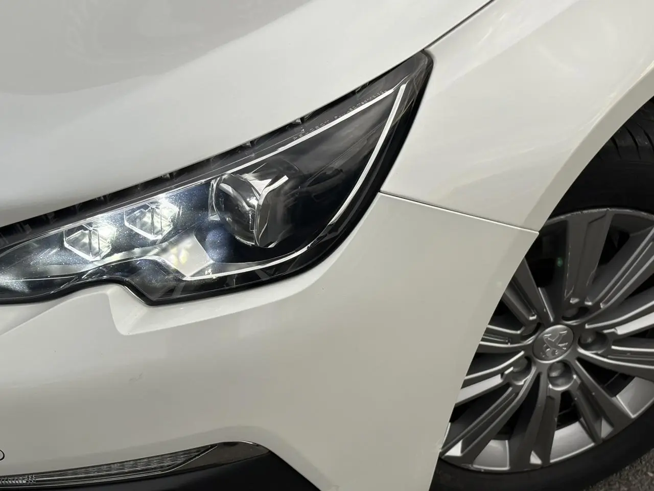 Gros plan sur le phare avant gauche et la jante alliage 16'' d'une Peugeot 308 blanc nacré de 2019.