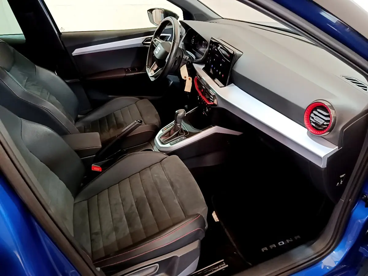 Intérieur avant droit du SEAT Arona bleu 2022, sièges noirs, volant cuir, tableau de bord avec écran tactile et aérateurs rouges.