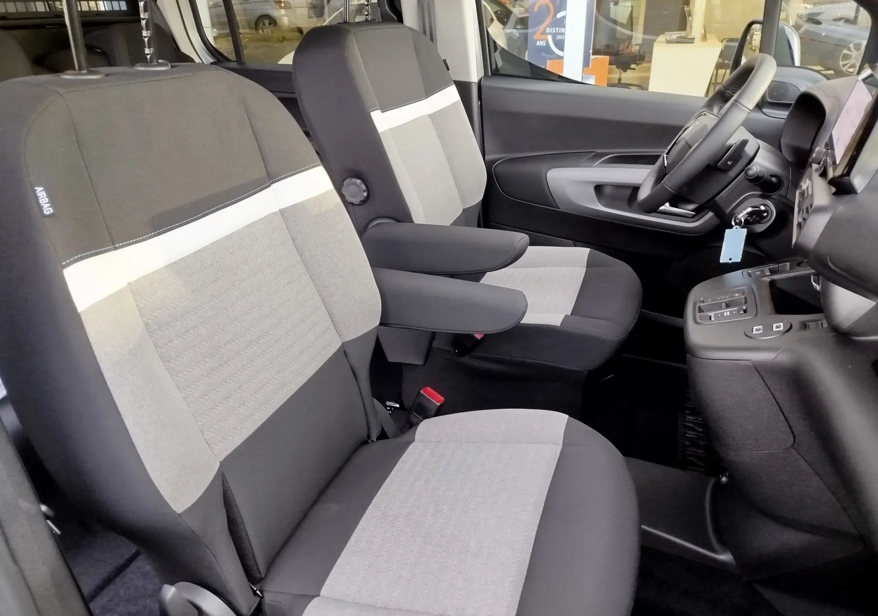 Vue intérieure côté conducteur du Citroën Berlingo 2026, sièges gris clair et foncé avec accoudoirs et volant visible.