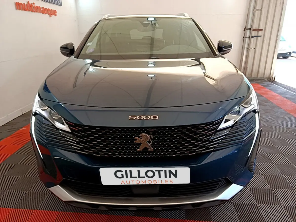 Vue frontale d'un Peugeot 5008 bleu avec calandre noire et logo lion, dans un showroom éclairé.