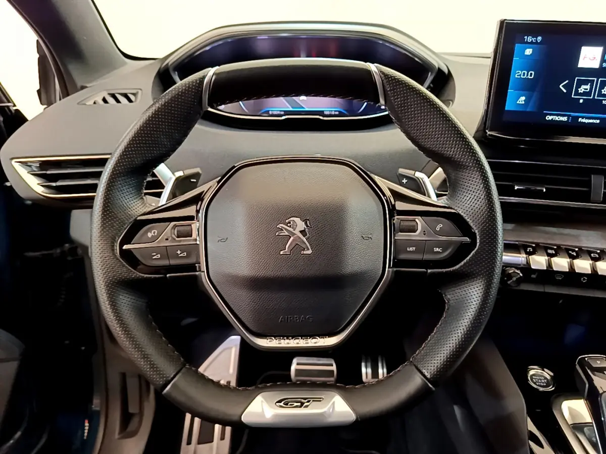 Vue rapprochée du volant cuir noir avec logo Peugeot et badge GT du SUV 5008 bleu, tableau de bord numérique visible.