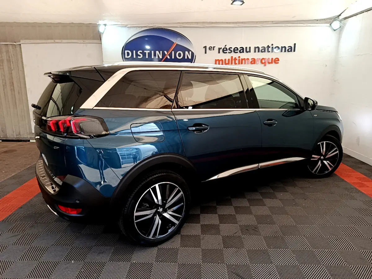 Peugeot 5008 bleu en 3/4 arrière droit, avec toit blanc et jantes alliage distinctives, en intérieur showroom.