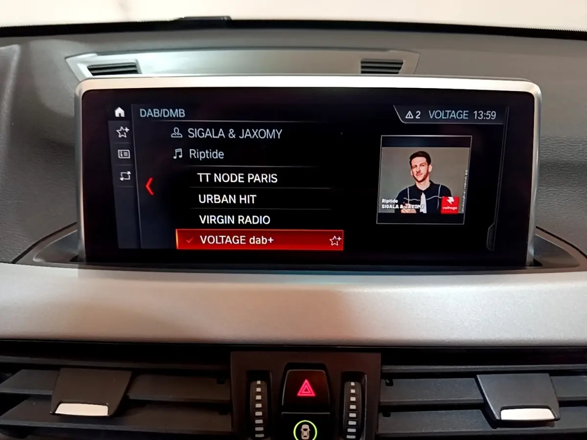 Écran tactile central du tableau de bord du BMW X1 noir, affichant la liste des radios DAB avec sélection sur Voltage dab+.