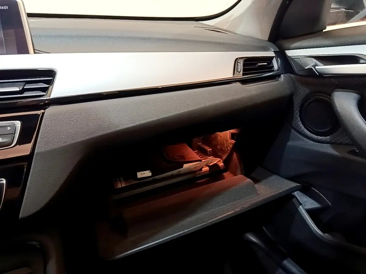 Gros plan sur la boîte à gants ouverte côté passager du BMW X1 noir, avec éclairage intérieur orange et tableau de bord noir.