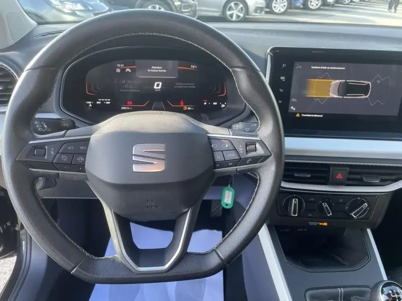 Vue intérieure centrée sur le volant noir du SEAT Arona 2023 avec tableau de bord numérique et écran central affichant l'aide au stationnement.
