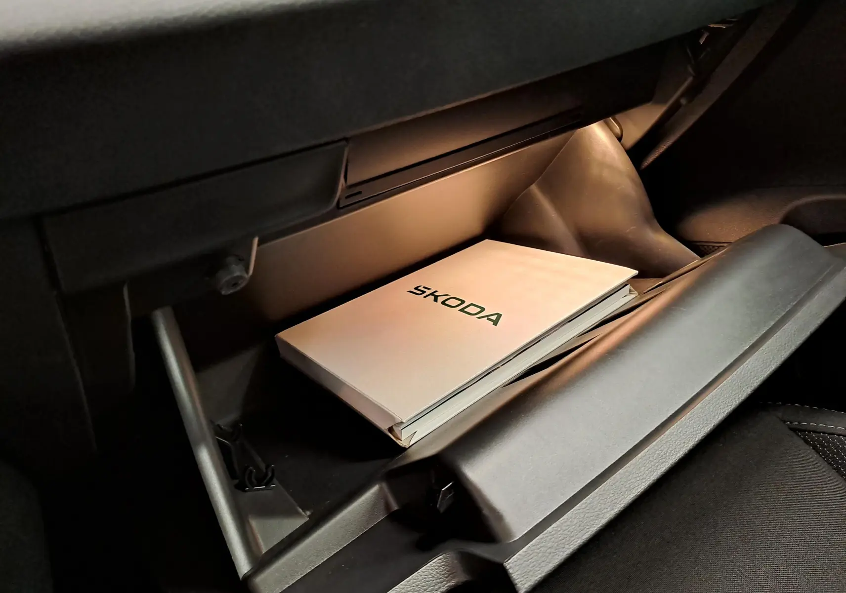 Gros plan sur la boîte à gants ouverte d’un Skoda Karoq 2025 blanc lune avec manuel d’utilisation visible à l’intérieur.